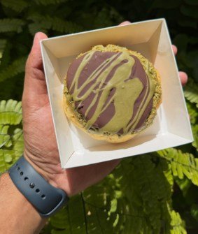 A round tart finished with chocolate and pistachio drizzle, boxed and held up.