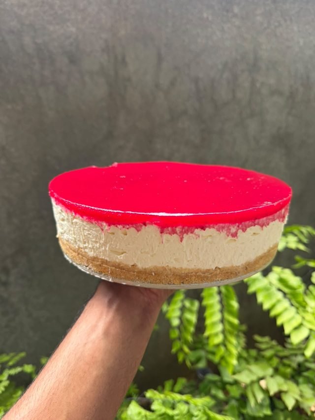 A cheesecake with a glossy ruby glaze over a biscuit base, held against a grey wall and ferns.